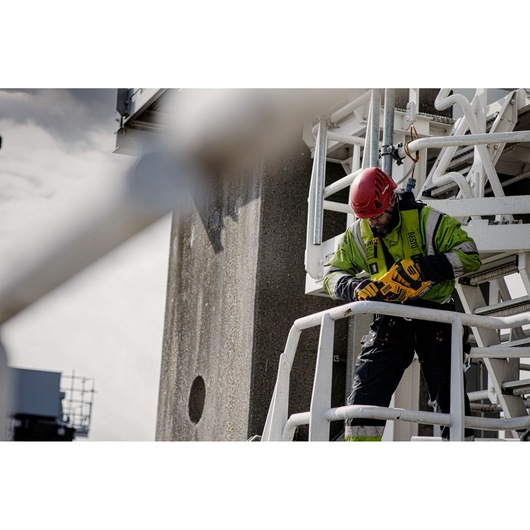 A construction worker wearing a safety helmet and high-visibility jacket is using a DEWALT power tool while standing on an industrial metal platform. The worker is secured with a safety harness.