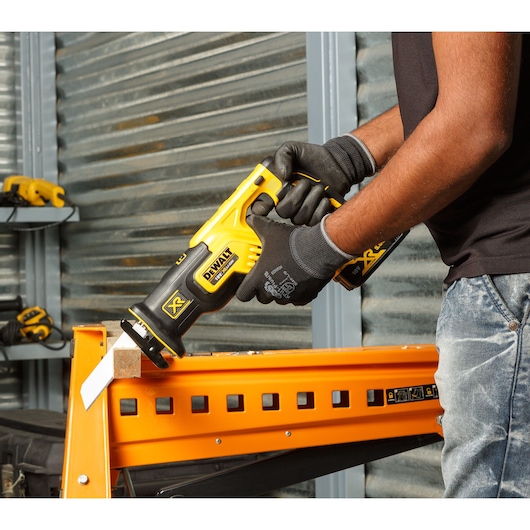 A person wearing black gloves is using a yellow and black DEWALT reciprocating saw to cut wood on an orange metal stand in a workshop.