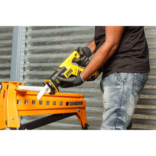 A person is using a DEWALT DCS367P2 cordless reciprocating saw to cut through a white pipe held on a yellow workbench. The person is wearing gloves and jeans, and the setting appears to be an industrial or workshop environment.