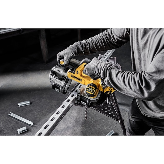 A person using a DEWALT cordless power tool to cut a metal beam, wearing gloves and a long-sleeved shirt, with the tool positioned on a metal stand and some metal pieces on the floor.