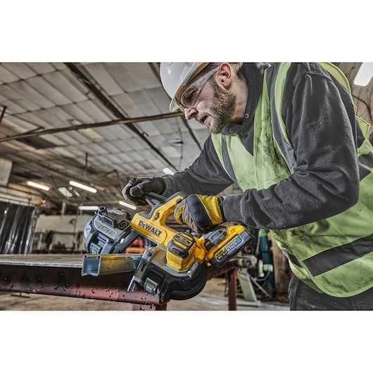 A person wearing a safety vest and gloves is operating a DEWALT cordless band saw (model DCS378N) to cut a metal beam in an industrial workshop.