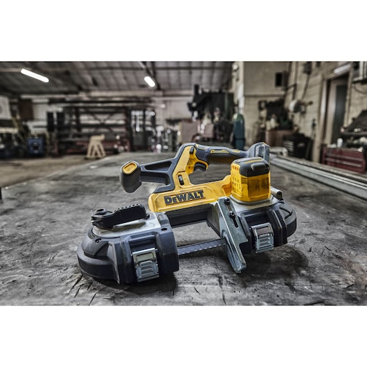 DEWALT cordless band saw (model DCS378N) placed on a workbench in an industrial workshop, with various tools and machinery in the background.