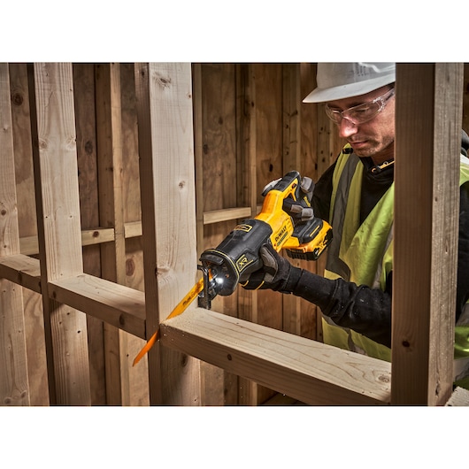 A person wearing a safety vest and hard hat uses a yellow and black DEWALT reciprocating saw to cut a wooden beam inside a construction site. The saw is battery-powered and fitted with a bright blade.