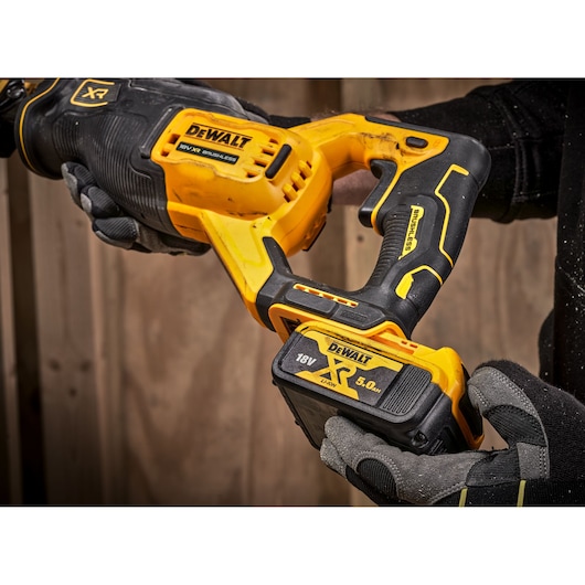 Close-up of a DEWALT 18V XR cordless reciprocating saw being held by gloved hands, with a visible 5.0Ah lithium-ion battery attached. The tool features yellow and black accents and is shown against a wooden background.