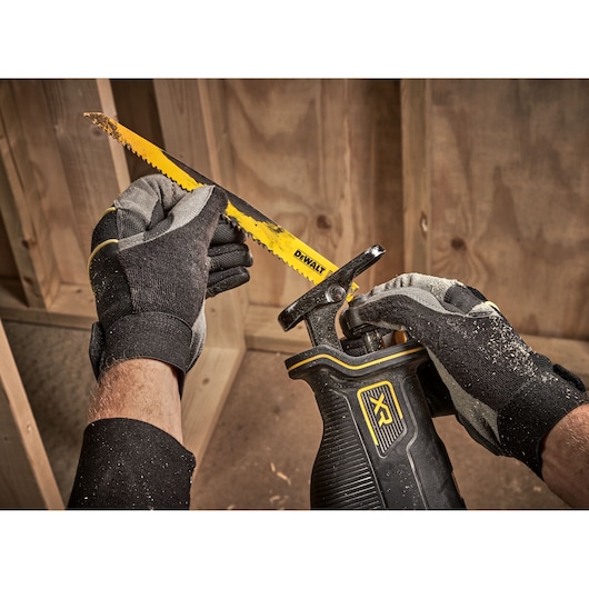 A close-up image of gloved hands installing a yellow DEWALT saw blade into a black and yellow reciprocating saw. The scene is set in a woodworking environment with wooden panels in the background.