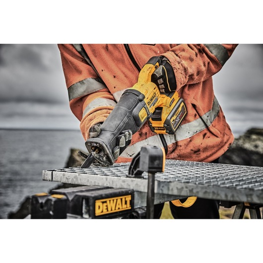 A person wearing an orange work jacket is using a DEWALT cordless reciprocating saw to cut through a metal grate outdoors. The DEWALT logo is visible on the saw and workbench.