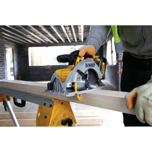 A person wearing gloves is using a DEWALT cordless circular saw to cut a wooden plank on a construction site.