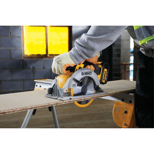 Person using DEWALT cordless circular saw to cut wood panel on workbench in workshop.