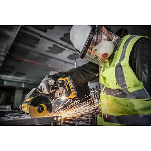 A person wearing protective gear, including a hard hat, face shield, mask, gloves, and a high-visibility vest, uses a DEWALT cordless power tool to cut metal, producing sparks.