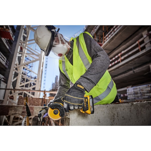 A construction worker wearing safety gear, including a helmet, face shield, gloves, and high-visibility vest, using a DEWALT DCS438E2T power tool to cut through metal at a building site.