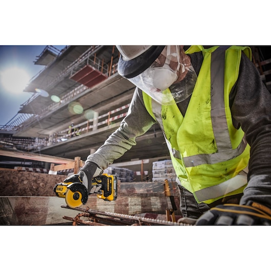 A construction worker wearing a yellow safety vest, gloves, face shield, and mask is using a DEWALT cordless rotary tool to cut metal rods at a construction site.