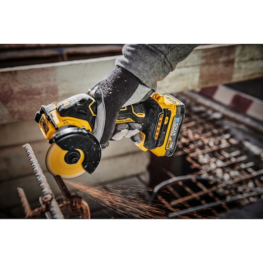 A person wearing gloves is using a DEWALT cordless brushless angle grinder to cut through a metal rod, producing sparks.