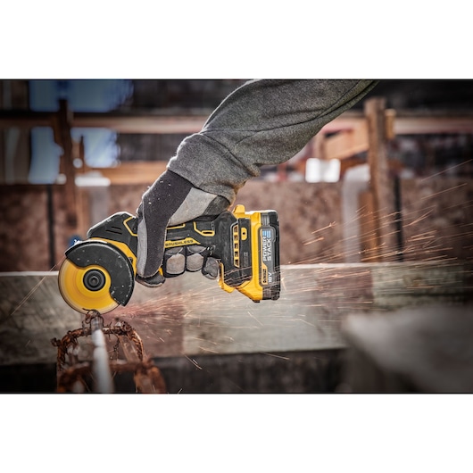 A person wearing protective gloves uses a DEWALT cordless brushless angle grinder to cut through a metal rod, with visible sparks flying from the point of contact.