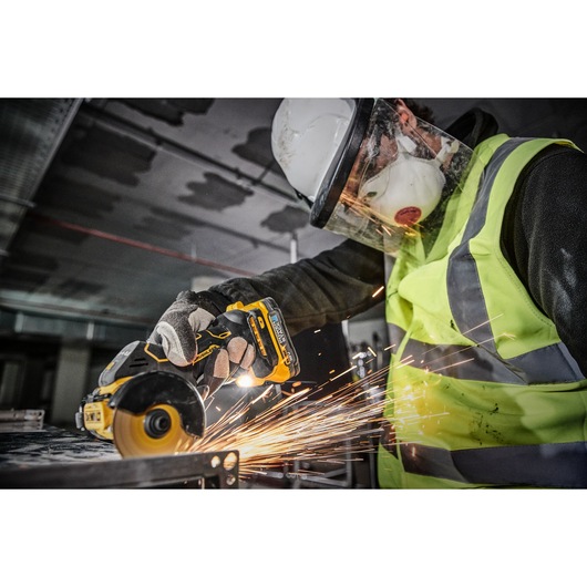 A person wearing safety gear, including a helmet, face shield, mask, gloves, and a high-visibility vest, is using a DEWALT DCS438N cordless power tool to cut metal, creating sparks.