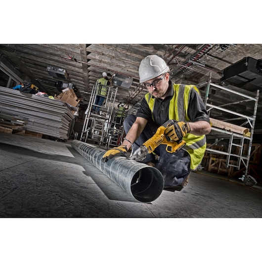 Worker uses DEWALT cordless metal shear on duct pipe at construction site. Safety gear and tools visible.
