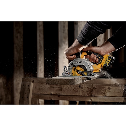 A person using a DEWALT cordless circular saw to cut a wooden plank on a workbench. Sawdust is visible as the tool operates.