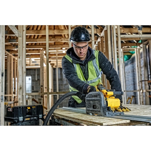 Worker uses DEWALT DCS520NT track saw to cut wood in a building under construction. Safety gear and tools are visible.