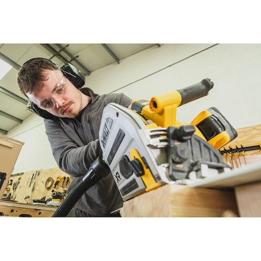 Person using a DEWALT cordless circular saw on wood, wearing safety glasses and ear protection in a workshop.