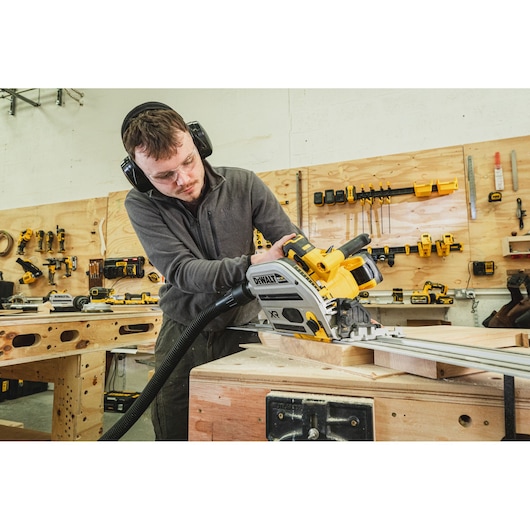 Person using a DEWALT cordless track saw on wood in a workshop, wearing ear and eye protection.