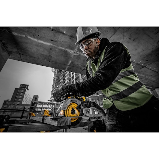 A construction worker wearing a safety vest and helmet operates a DEWALT circular saw (model DCS565NT) to cut a wooden plank at a building site. The background shows concrete structures and high-rise buildings under construction.