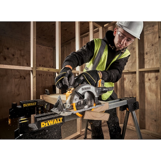 A person wearing safety gear uses a DEWALT cordless circular saw (model DCS565N) to cut wood on a sawhorse at a construction site.