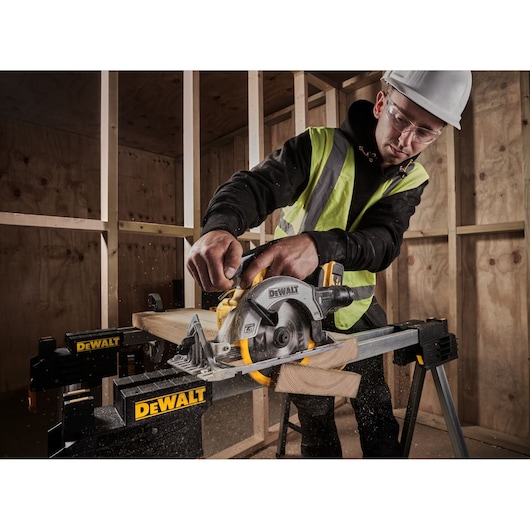 A person wearing a safety helmet and high-visibility vest uses a DEWALT DCS565N cordless circular saw to cut a wooden plank on a DEWALT workbench in a construction site.