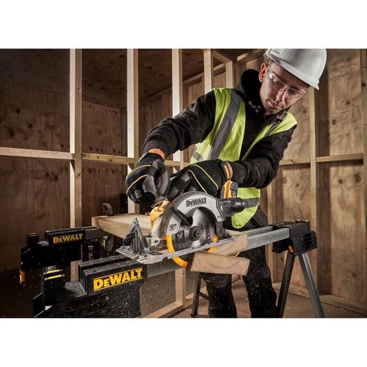 A person wearing safety gear uses a DEWALT DCS565P2 cordless circular saw to cut wood on a DEWALT workbench inside a construction site with wooden framing.