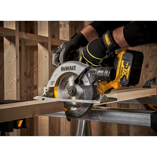 A DEWALT cordless circular saw being used by a person wearing gloves to cut a wooden plank in a workshop setting.