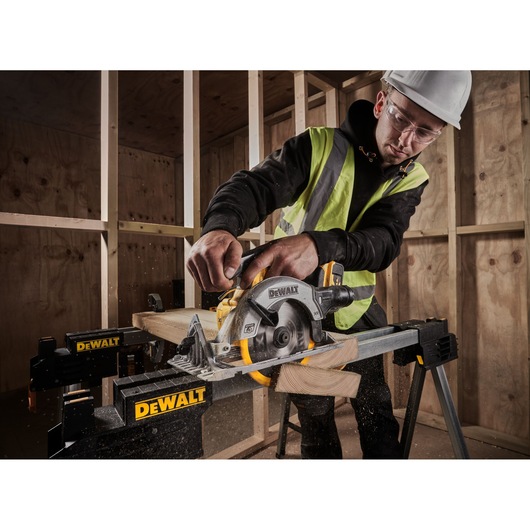 A person wearing a safety vest and helmet is using a DEWALT cordless circular saw (model DCS565P2) to cut wood on a DEWALT workbench inside a partially constructed wooden structure.