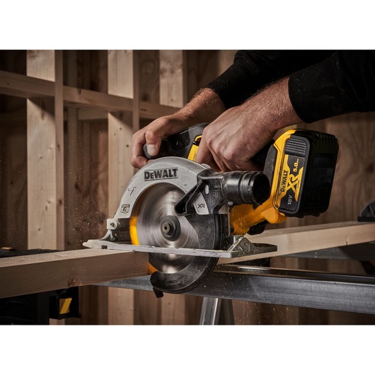 A person using a DEWALT cordless circular saw to cut a wooden plank in a workshop setting.