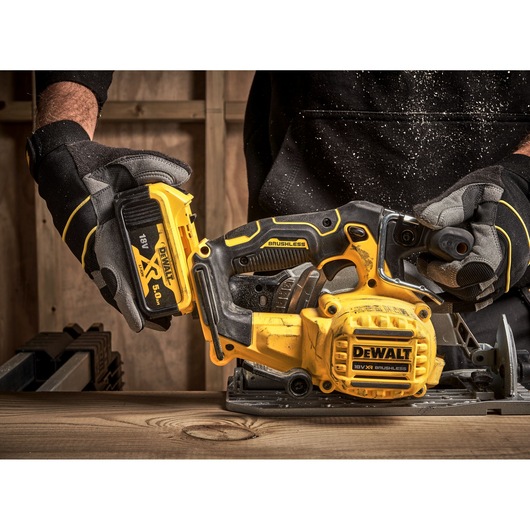 A person wearing gloves is holding a yellow and black DEWALT cordless brushless circular saw, model DCS565P2, and inserting an 18V XR 5.0Ah battery. The saw is placed on a wooden surface with sawdust around.