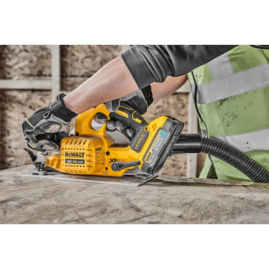 A person wearing safety gloves and a high-visibility vest is using a DEWALT cordless circular saw with a dust extraction hose attached, cutting wood on a construction site.