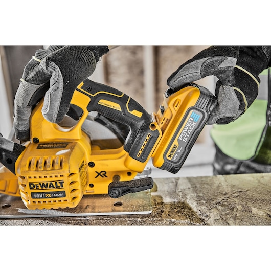 A close-up image of a person wearing work gloves attaching an 18V DEWALT POWERSTACK XR Li-Ion battery to a DEWALT brushless cordless circular saw on a workbench.