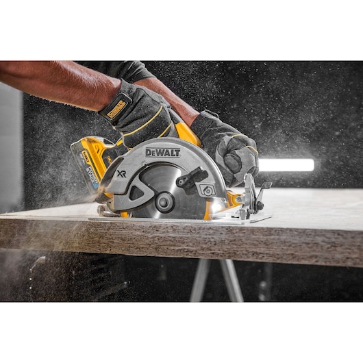A close-up of a person using a DEWALT cordless circular saw to cut through a piece of wood, with sawdust flying in the air. The person is wearing protective gloves.