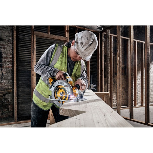 A person wearing a safety vest and hard hat using a DEWALT DCS573H2T cordless circular saw to cut a large wooden plank in an unfinished construction site.