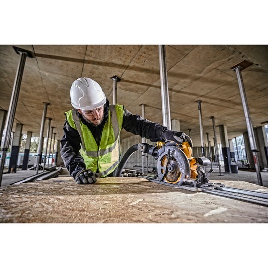 Worker using DEWALT DCS579T2 circular saw on wood panel at a construction site, wearing safety gear.