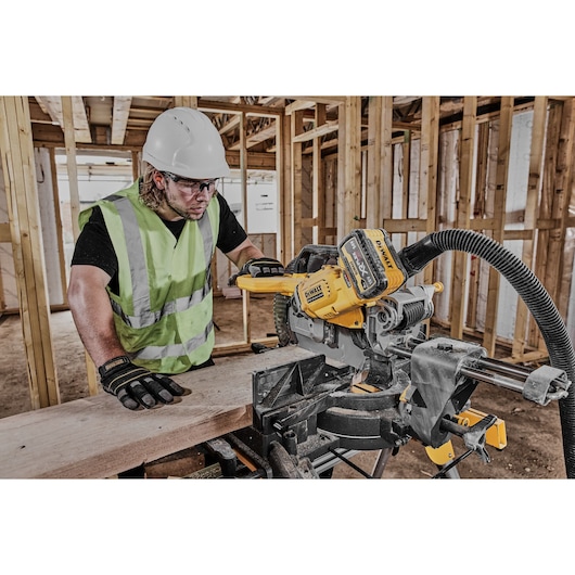 A person wearing a reflective safety vest, hard hat, and gloves is operating a DEWALT cordless miter saw (model DCS781N) on a construction site with wooden frames.