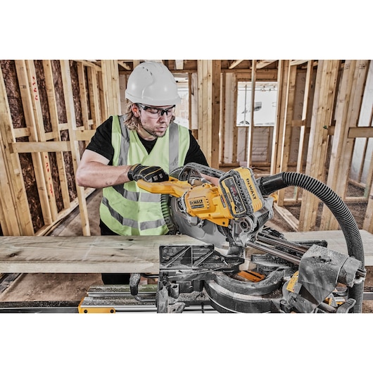 A construction worker, wearing a high-visibility vest and white hard hat, is using a DEWALT DCS781N cordless mitre saw to cut a wooden plank at a building site framed with timber.