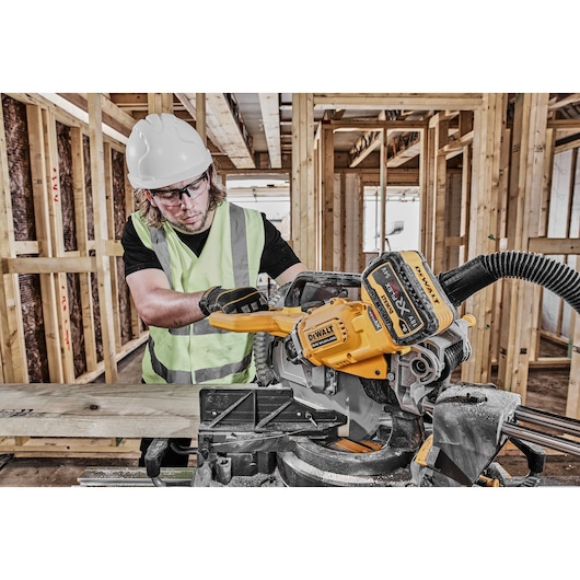 A person wearing a safety helmet and high-visibility vest uses a DEWALT DCS781N power saw to cut a piece of wood in a construction site with wooden framing.