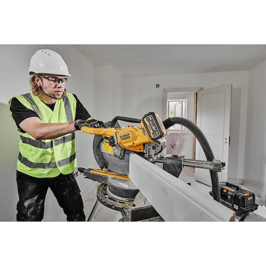 A person wearing a safety helmet and reflective vest operates a DEWALT DCS781N cordless miter saw, cutting a piece of wood in a construction or renovation setting.