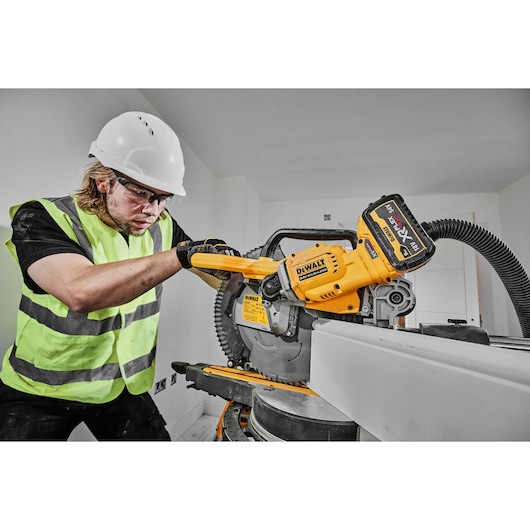 A person wearing a high-visibility vest and a hard hat is operating a DEWALT DCS781N power tool, specifically a cordless miter saw, in an indoor construction setting.