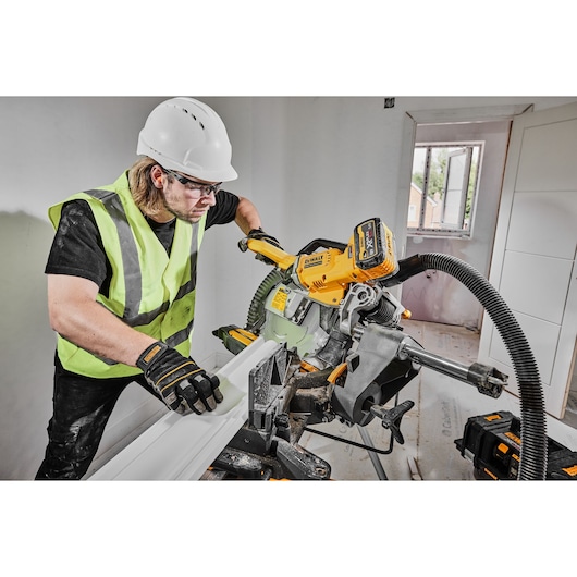 A construction worker wearing a safety helmet, high-visibility vest, and gloves is operating a DEWALT DCS781N cordless mitre saw indoors, cutting a piece of white material. The saw is attached to a dust extraction hose.
