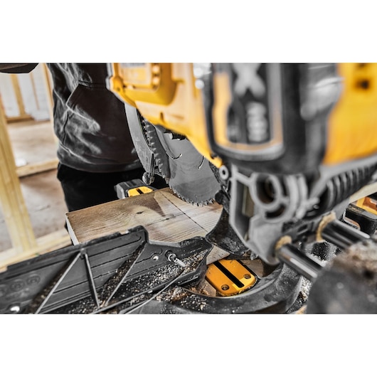 Close-up of a DEWALT DCS781N miter saw cutting a wooden plank in a workshop setting. Sawdust is visible around the saw and plank.