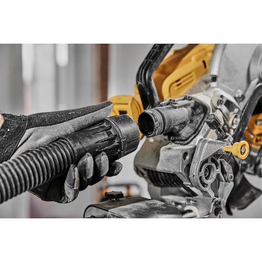 Close-up of a person wearing gloves connecting a dust extraction hose to a DEWALT power tool, likely a saw, in a workshop setting.