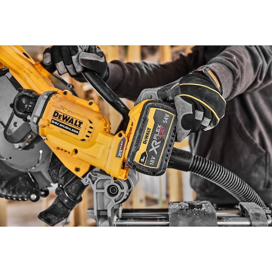 A person wearing protective gloves installs a DEWALT XR FLEXVOLT 18V/54V rechargeable battery onto a yellow DEWALT saw brushless power tool, in a woodworking or construction setting.