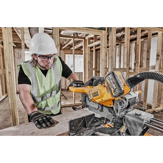 A construction worker wearing a safety helmet, high-visibility vest, and gloves is using a DEWALT DCS781X2 cordless power tool with an XR battery for cutting wood inside a partially built wooden structure.