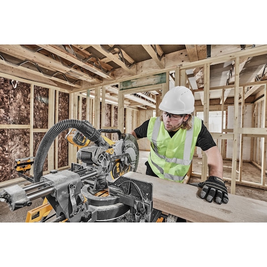 A person wearing a white hard hat, reflective safety vest, and gloves is using a DEWALT miter saw to cut a piece of wood inside a building under construction. The saw is equipped with a dust extraction hose and the surroundings show exposed wooden framing.