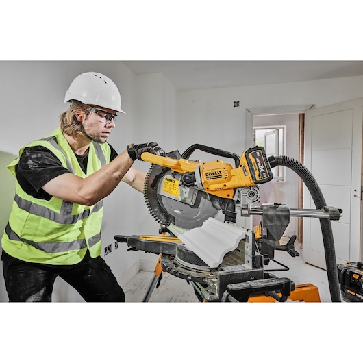 A person wearing a safety helmet and high-visibility vest operates a DEWALT DCS781X2 sliding compound miter saw in a room under construction. The saw is cutting a piece of white trim, and a vacuum hose is attached for dust extraction.