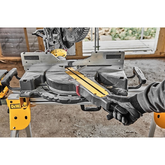 A DEWALT sliding compound miter saw with a yellow and black design is being operated by a person wearing gloves. The saw is set up on a sturdy yellow stand in a workshop environment.