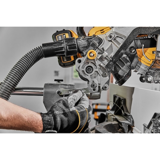 Close-up of a person wearing work gloves adjusting a part on a DEWALT cordless miter saw with dust extraction hose attached.